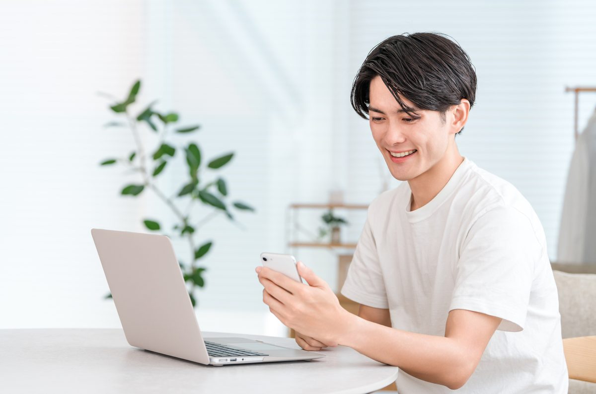 自宅のテーブルでノートパソコンとスマートフォンを見ながら作業する男性